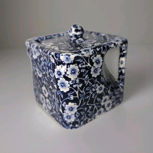 Puede incluir: Contenedor cuadrado de cerámica con tapa, con un estampado floral azul y blanco. Tiene un pequeño asa y un pomo en la tapa. El diseño es de estilo clásico y vintage.