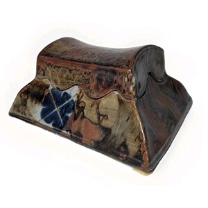 May include: A rectangular ceramic box with a curved, saddle-shaped lid. The piece features a glossy, multi-toned glaze in shades of brown, tan, and black, with a distinct blue four-leaf clover pattern visible on the front side of the base.