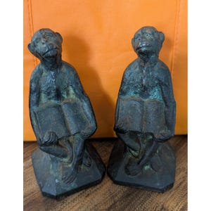 May include: Two antique bronze monkey bookends. Each monkey is seated and holding a book. The bookends have a dark, aged patina, and are set on a hexagonal base. The background is a solid orange color.