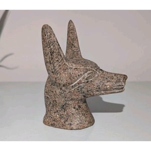 Puede incluir: Una escultura detallada de la cabeza de un chacal, probablemente representando a Anubis, el antiguo dios egipcio del más allá. La escultura está hecha de una piedra moteada de color marrón rojizo, con detalles tallados que definen los rasgos. Las orejas son erguidas y puntiagudas.