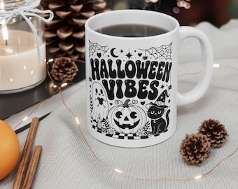 May include: A white ceramic mug featuring a black graphic print with the text HALLOWEEN VIBES. The design includes a ghost, a jack-o'-lantern, a cat wearing a witch hat, spiderwebs, stars, and hearts. The mug has a handle and a cylindrical shape.