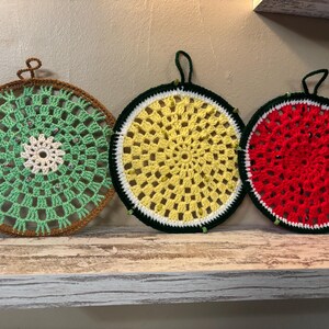 May include: Three round, crocheted pot holders in a row. One is green with a brown border, another is yellow with a black border, and the last is red with a green border. Each has a loop for hanging and decorative beads.