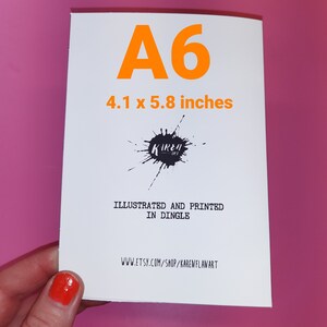 May include: A white card with the text "A6 4.1 x 5.8 inches" printed in orange. The card also has a black ink splatter with the text "Kireh" in white. The text "Illustrated and printed in Dingle" and "www.etsy.com/shop/karewflanart" are also printed on the card.