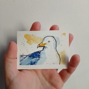 Könnte beinhalten: Ein kleines rechteckiges Aquarellbild auf strukturiertem Papier, das eine Möwe mit weißem Kopf, gelbem Schnabel mit roter Spitze und blauen Flügeln zeigt. Der Hintergrund besteht aus sanften gelben und blauen Aquarellfarben mit kleinen Spritzern.