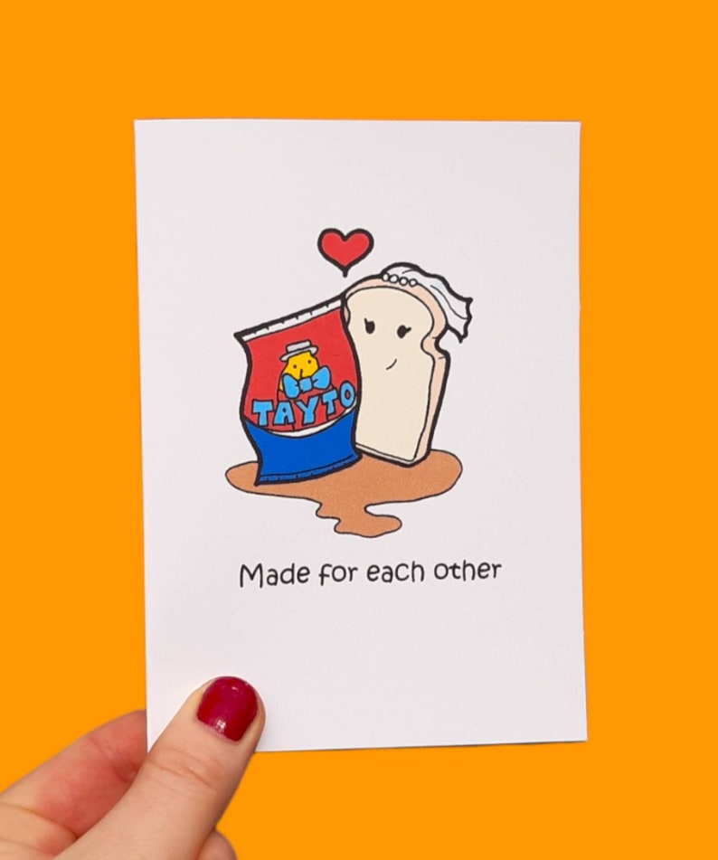 Peut inclure: Une carte de v&oelig;ux avec une illustration de dessin anim&eacute; d'un sac de chips Tayto et une tranche de pain. Les chips et le pain sont dessin&eacute;s avec des visages et le pain porte un voile. Le texte "Made for each other" est imprim&eacute; sous l'illustration.