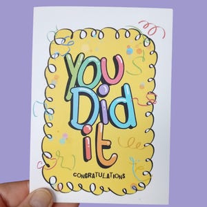 May include: A rectangular greeting card with a yellow centre featuring the text "You Did it" in large, colourful bubble letters. Below the main text, the word "CONGRATULATIONS" is printed in black. The design includes a black squiggly border.