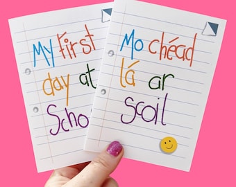 May include: Two white paper notes with colorful text that reads "My first day at School" and "Mo chéad lá ar scoil". A yellow smiley face sticker is on one note. The background is bright pink.