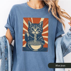 May include: Blue jean colored t-shirt featuring a graphic of a cat eating noodles with chopsticks from a bowl. The cat is set against a red and cream sunburst design. The shirt is a comfortable, casual style.