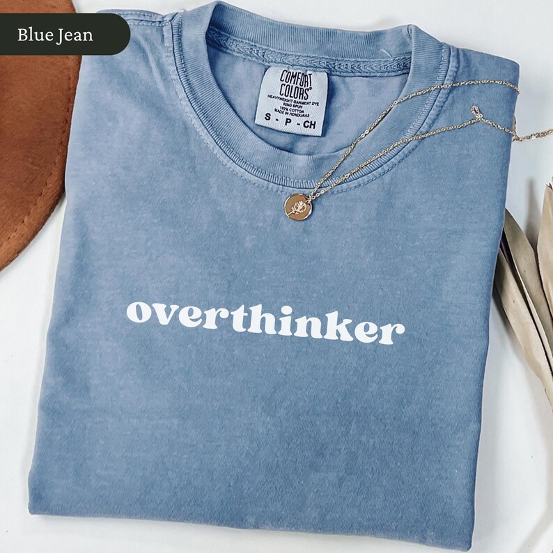 May include: A blue jean coloured t-shirt with the word "overthinker" printed in white. A gold necklace with a small pendant rests on the shirt. The shirt has a Comfort Colors tag.