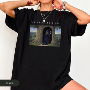 Op de afbeelding: Zwart T-shirt met de tekst "I'LL BE IN MY OFFICE" boven een fantasie-illustratie. De illustratie toont een tovenaar in een donkerblauwe jurk in een boomdeur, met een gloeiende bol.