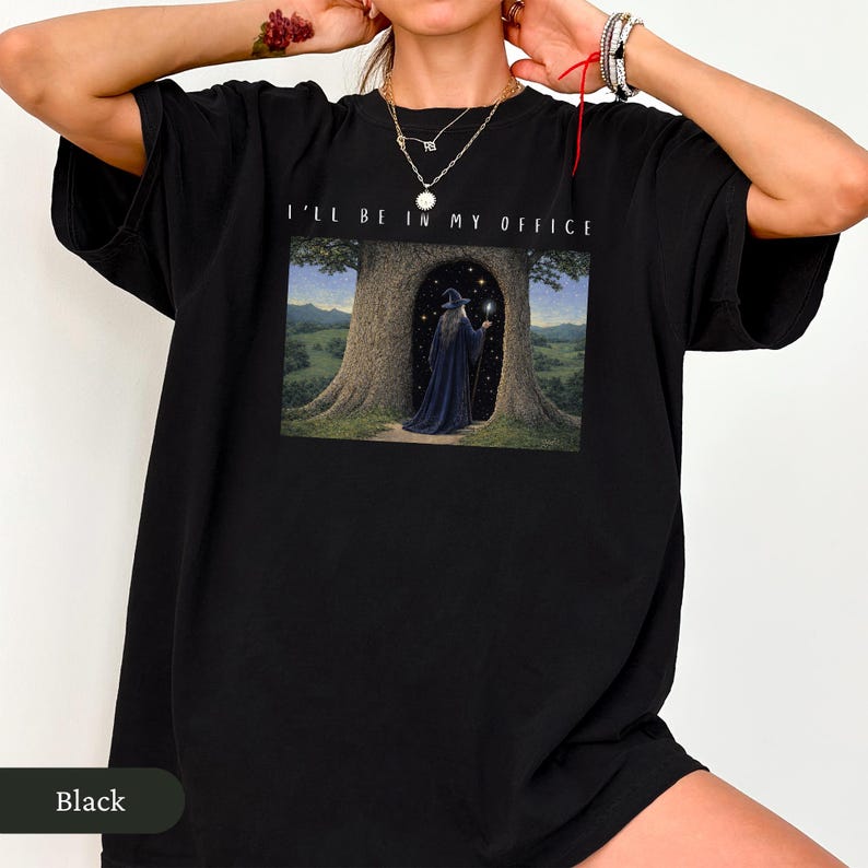 Op de afbeelding: Zwart T-shirt met de tekst "I'LL BE IN MY OFFICE" boven een fantasie-illustratie. De illustratie toont een tovenaar in een donkerblauwe jurk in een boomdeur, met een gloeiende bol.
