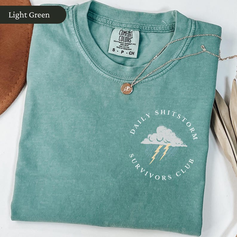 May include: Light green t-shirt with the text "DAILY SHITSTORM SURVIVORS CLUB" in a circular design. The design includes a cloud, lightning bolts, and text. A gold necklace with a pendant rests on the shirt.