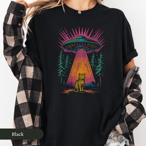 Op de afbeelding: Zwart T-shirt met een kleurrijke afbeelding van een UFO die een kat beschijnt. Het ontwerp bevat een boslandschap met een lichtstraal. Het woord "Black" staat onderaan.