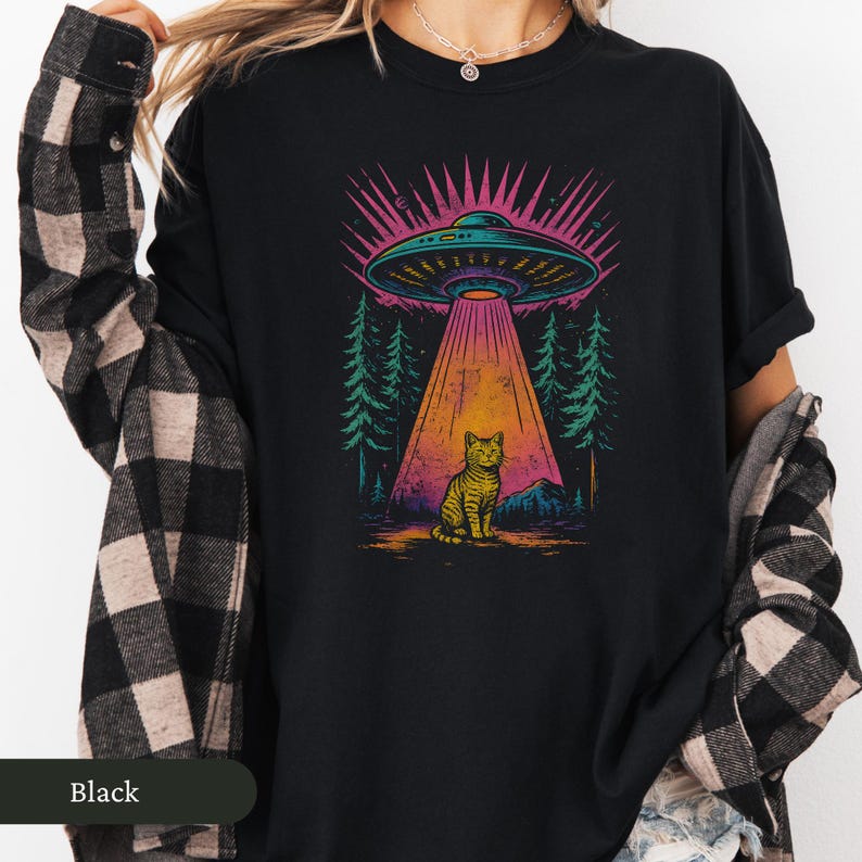 Op de afbeelding: Zwart T-shirt met een kleurrijke afbeelding van een UFO die een kat beschijnt. Het ontwerp bevat een boslandschap met een lichtstraal. Het woord "Black" staat onderaan.