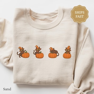 May include: A sand-coloured sweatshirt with a crew neck, displaying a design of four black cats in witch hats on orange pumpkins. The graphic is centered. The text "Sand" is at the bottom left. The top has long sleeves.