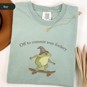 Puede incluir: Camiseta verde claro con la frase "Off to commit tom foolery" y una rana de dibujos animados en monopat&iacute;n, con un sombrero de mago. Un collar dorado est&aacute; colocado sobre la camiseta.