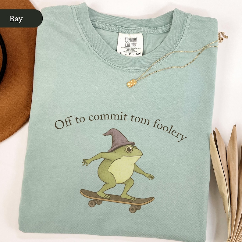 Puede incluir: Camiseta verde claro con la frase "Off to commit tom foolery" y una rana de dibujos animados en monopat&iacute;n, con un sombrero de mago. Un collar dorado est&aacute; colocado sobre la camiseta.