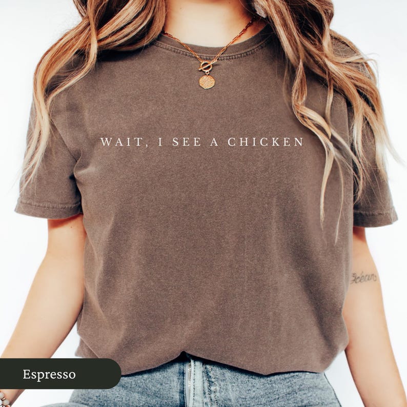 May include: Espresso-coloured t-shirt with the text "WAIT, I SEE A CHICKEN" in white. The shirt is a short-sleeved crew neck style. The model is wearing a gold necklace.