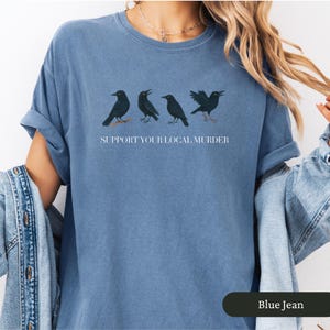 May include: Blue jean-colored t-shirt with a graphic of four black crows and the text "SUPPORT YOUR LOCAL MURDER". The shirt has a relaxed fit and rolled-up sleeves. The bottom right corner of the shirt has the text "Blue Jean".