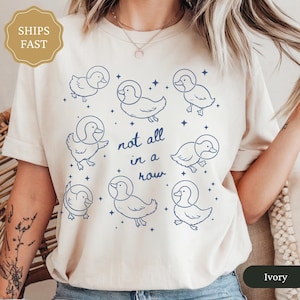 May include: Ivory-colored t-shirt featuring a whimsical design of cartoon ducks wearing space helmets, with the phrase "not all in a row" in blue script. The ducks are surrounded by small star graphics.