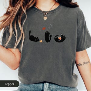 May include: Dark gray t-shirt featuring three black cat illustrations. One cat is playing with a leaf, another is standing, and the third is rolling on its back with a leaf. The word "Pepper" is at the bottom of the image.
