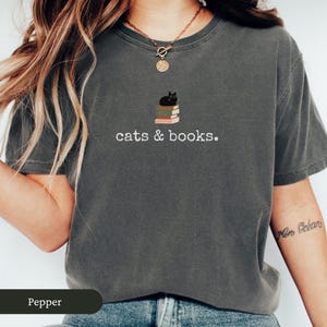 Może przedstawiać: Szary t-shirt z nadrukiem czarnego kota siedzącego na stosie książek. Na koszulce widnieje napis "cats & books."