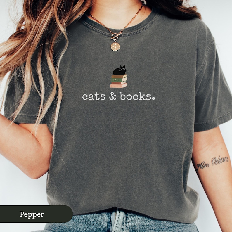 Może przedstawiać: Szary t-shirt z nadrukiem czarnego kota siedzącego na stosie książek. Na koszulce widnieje napis "cats & books."
