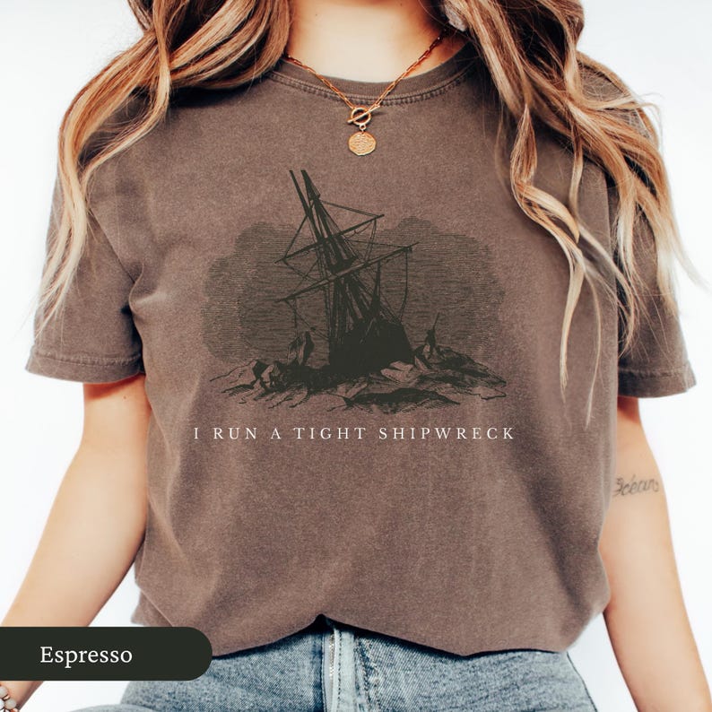May include: Espresso-colored t-shirt featuring a distressed graphic of a shipwreck. The text "I RUN A TIGHT SHIPWRECK" is printed below the image. The shirt has short sleeves and a crew neck.