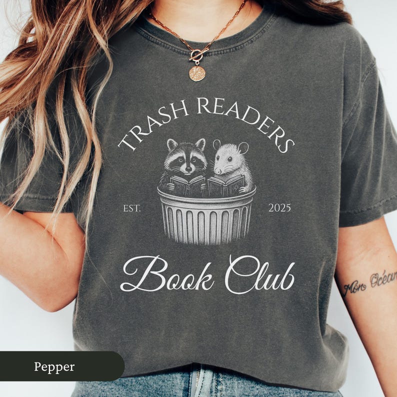 Puede incluir: Una camiseta gris oscuro con el texto "Trash Readers Book Club" en blanco. El texto est&aacute; rodeado de un c&iacute;rculo con las palabras "Trash Readers" en la parte superior y "Est. 2025" en la parte inferior. Hay una ilustraci&oacute;n de un mapache y una rata leyendo libros en un cubo de basura.