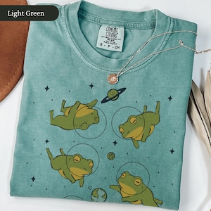 May include: Light green Comfort Colors t-shirt featuring a graphic of four cartoon frogs in space suits, with planets and stars. A silver necklace with a pendant is draped across the shirt. The shirt is folded neatly.