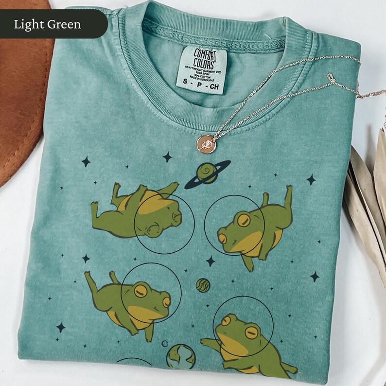 May include: Light green Comfort Colors t-shirt featuring a graphic of four cartoon frogs in space suits, with planets and stars. A silver necklace with a pendant is draped across the shirt. The shirt is folded neatly.