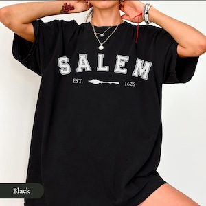 Peut inclure: T-shirt noir avec le mot "SALEM" en lettres blanches, avec "EST. 1626" et un dessin de balai en dessous. Le t-shirt est de style d&eacute;contract&eacute;, adapt&eacute; au quotidien.