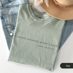 Puede incluir: Camiseta verde claro con la cita "I should infinitely prefer a book. - Jane Austen" en texto negro. La camiseta est&aacute; doblada sobre unos vaqueros azules, con un sombrero beige y un collar plateado.