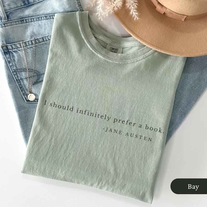 Puede incluir: Camiseta verde claro con la cita "I should infinitely prefer a book. - Jane Austen" en texto negro. La camiseta est&aacute; doblada sobre unos vaqueros azules, con un sombrero beige y un collar plateado.