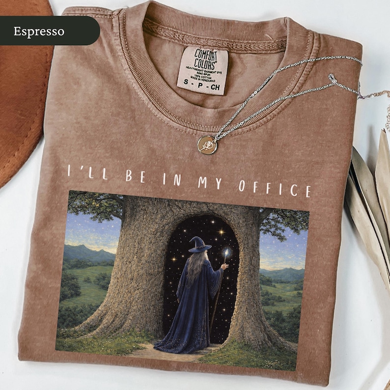 Op de afbeelding: Espresso-kleurig T-shirt met de tekst "I'll Be In My Office" boven een afbeelding van een tovenaar. Het shirt heeft een ronde hals en korte mouwen. Een zilveren ketting met een hanger ligt op het shirt.