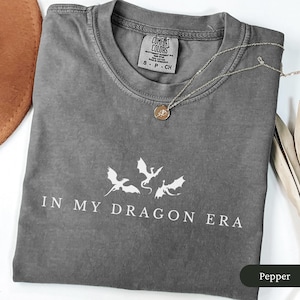 May include: A grey t-shirt with the text "IN MY DRAGON ERA" and a graphic of three dragons. The shirt is folded and has a Comfort Colors tag. A silver necklace with a pendant rests on the shirt.