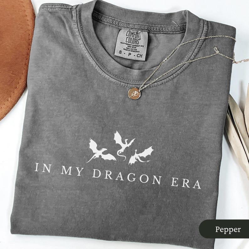 May include: A gray t-shirt with the text "IN MY DRAGON ERA" and a graphic of three dragons. The shirt is folded and has a Comfort Colors tag. A silver necklace with a pendant rests on the shirt.