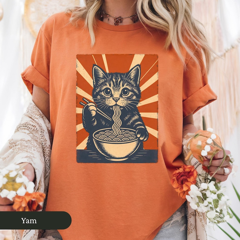 May include: Orange t-shirt featuring a graphic of a cat eating ramen noodles with chopsticks. The cat is black and gray, and the ramen bowl is blue. The background has a retro sunburst design. The word "Yam" is at the bottom.