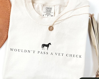 May include: Ivory-colored t-shirt with the text "WOULDN'T PASS A VET CHECK" and a black horse silhouette. A gold-colored necklace with a round pendant is draped over the shirt. The shirt is a Comfort Colors brand, size small.