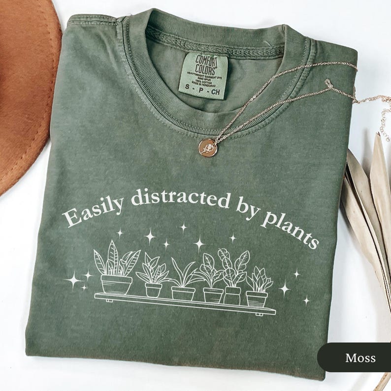 May include: A moss green t-shirt featuring the phrase "Easily distracted by plants" and a plant graphic. A gold necklace with a pendant is on the shirt. The t-shirt is folded on a white surface.
