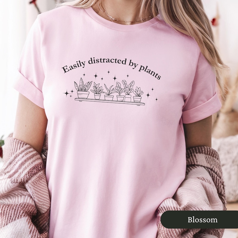 May include: A pale pink t-shirt featuring the phrase "Easily distracted by plants" above a shelf of potted plants. The design is in black and white. The shirt has short sleeves and a round neck.