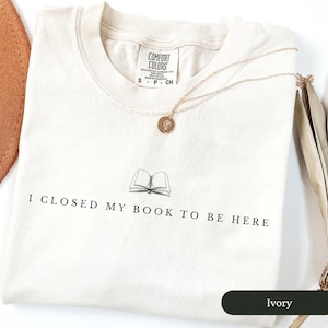 May include: Ivory-colored t-shirt with the text "I CLOSED MY BOOK TO BE HERE" and a graphic of an open book. A gold-colored necklace with a round pendant rests on the shirt. The shirt is labeled "Comfort Colors".