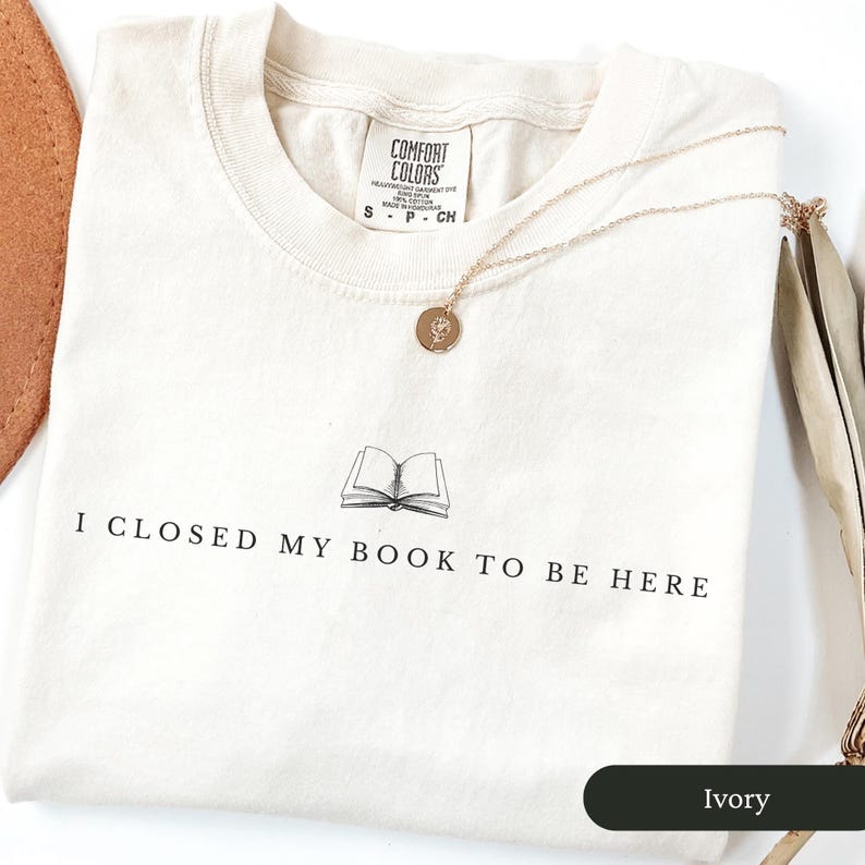 May include: Ivory-colored t-shirt with the text "I CLOSED MY BOOK TO BE HERE" and a graphic of an open book. A gold-colored necklace with a round pendant rests on the shirt. The shirt is labeled "Comfort Colors".