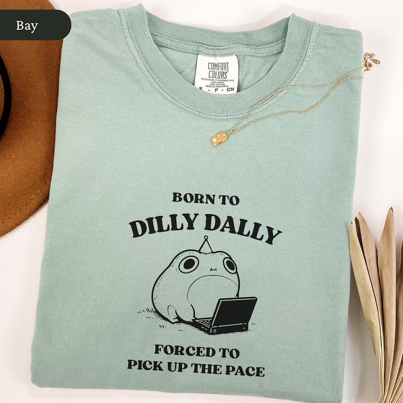 May include: A light green Comfort Colors t-shirt with the text "BORN TO DILLY DALLY FORCED TO PICK UP THE PACE" and a cartoon frog with a laptop. A gold necklace is draped across the shirt.