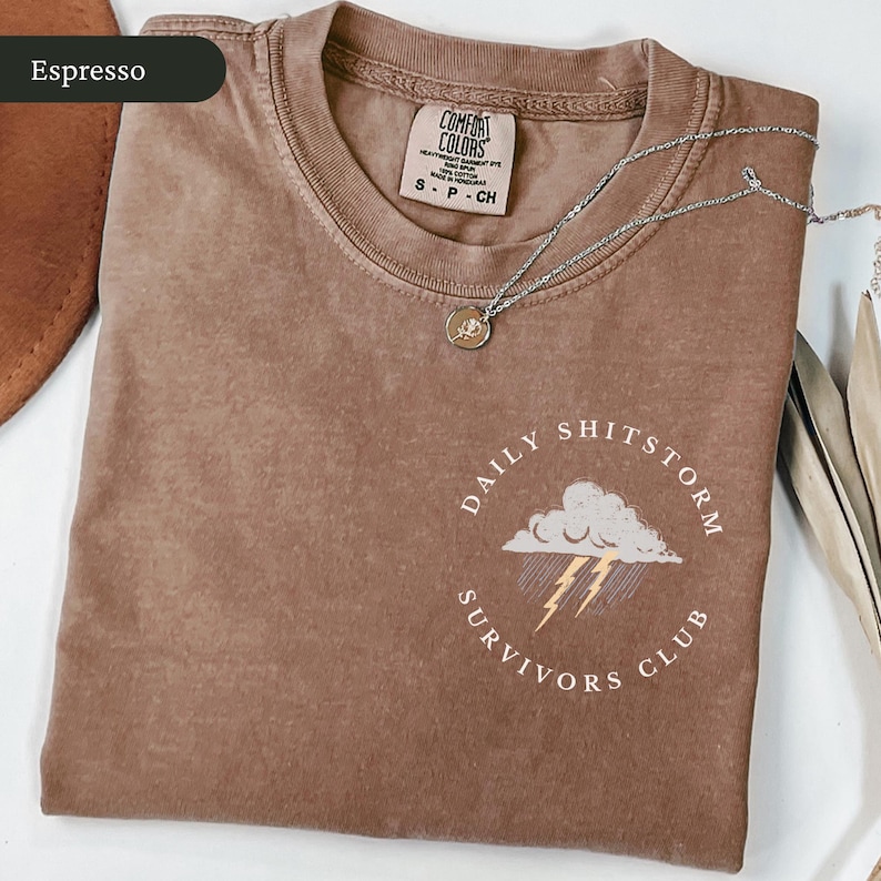 May include: Espresso-colored Comfort Colors t-shirt with a crew neck. The shirt features a graphic of a cloud with lightning and the text "DAILY SHITSTORM SURVIVORS CLUB". A silver necklace with a pendant is on the shirt.