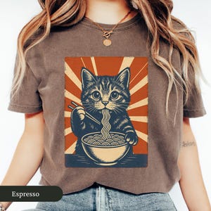 May include: Espresso-colored t-shirt featuring a graphic of a cat eating ramen noodles. The cat illustration is in a vintage style with a red and cream background. The text "Espresso" is at the bottom of the shirt.