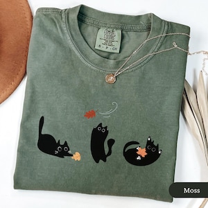 May include: A moss green Comfort Colors t-shirt featuring three black cat illustrations playing with autumn leaves. A gold necklace with a pendant is draped over the shirt. The shirt has a crew neck and short sleeves.