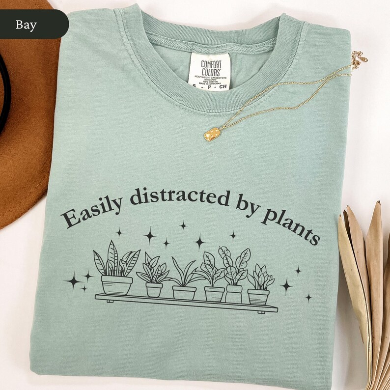 May include: A pale green t-shirt featuring the text "Easily distracted by plants" and a plant graphic. A gold necklace with a pendant is on the shirt. The shirt has a Comfort Colors label.