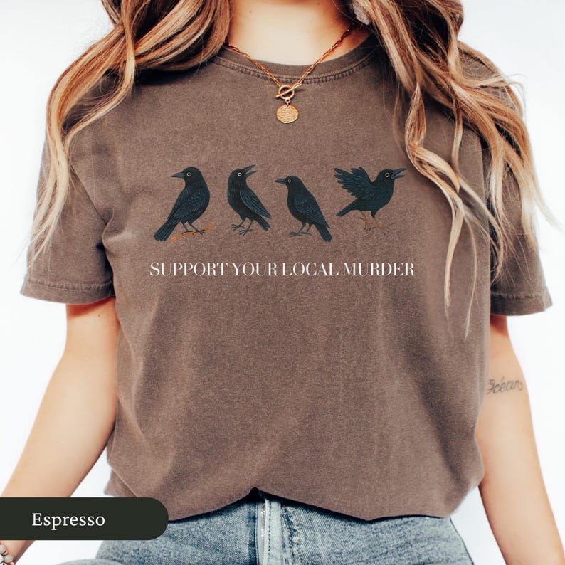 May include: Espresso-colored t-shirt featuring four black crow illustrations and the text "SUPPORT YOUR LOCAL MURDER". The crows are depicted in various poses. The shirt is a comfortable, casual wear item.