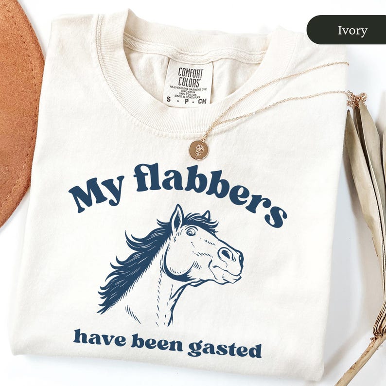 May include: Ivory-colored t-shirt with the phrase "My flabbers have been gasted" in navy blue, along with a horse illustration. A gold necklace with a pendant is draped over the shirt. The shirt is made of a soft material.